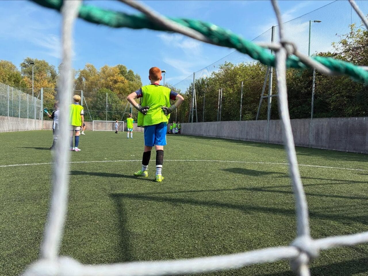 View through the gap of a football of young goalkeeper and other children on the pitch.
