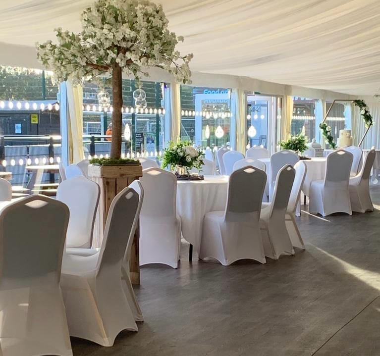 Elegant banqueting setup inside the draped marquee at SSFC, showing round tables with white linens, white-covered chairs, and a decorative center tree with hanging glass baubles, with large glass doors looking out onto the grounds.
